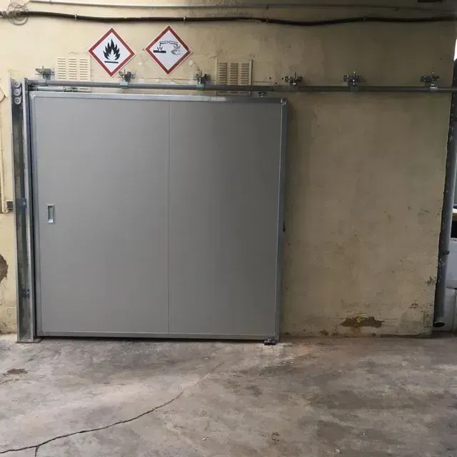 Puerta corrediza gris en un entorno industrial de hormigón, con señales de peligro encima.