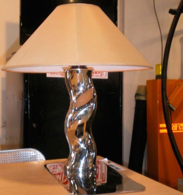 Lámpara cromada con pantalla color crema sobre mesa, reflejando los objetos circundantes.