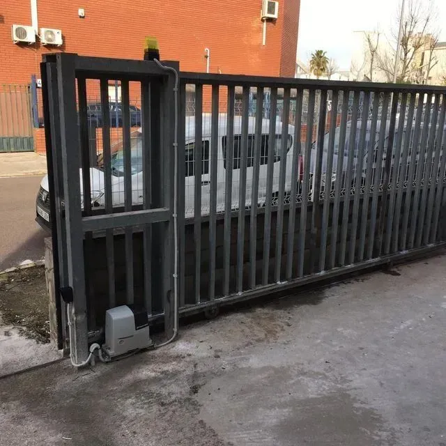 Puerta corrediza de metal de color gris con barras verticales, motor eléctrico y luz intermitente incorporada.
