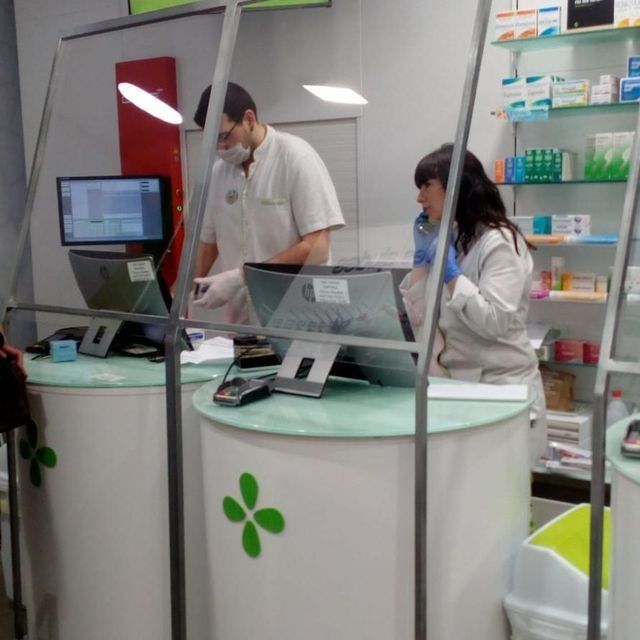 Dos empleados de farmacia detrás de una barrera protectora en un mostrador, con máscaras y guantes.