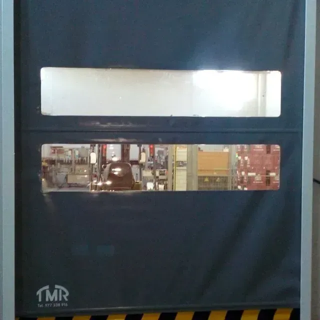 Puerta industrial gris con dos ventanas que muestra el interior de una fábrica. La puerta tiene franjas de advertencia.