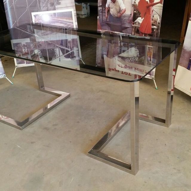 Mesa con tapa de cristal y patas de acero inoxidable en forma de Z; configuración interior, varios carteles de fondo.