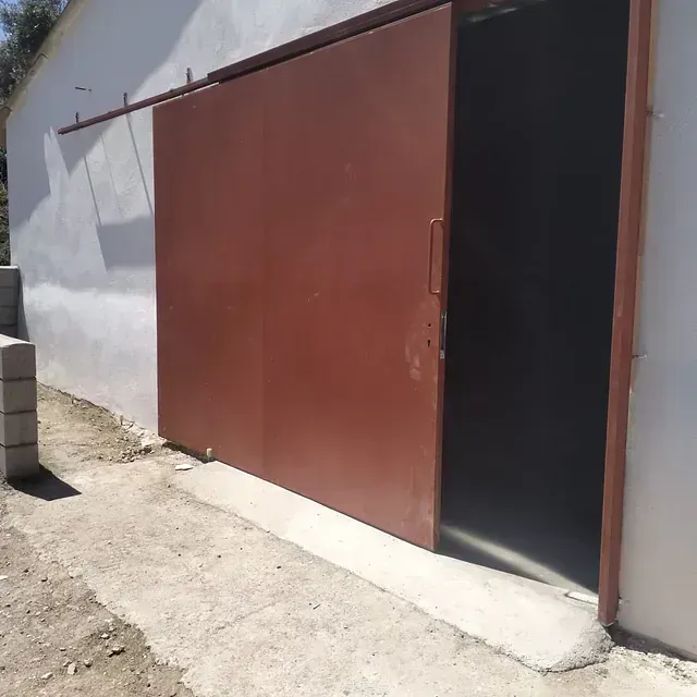 Puerta corrediza metálica de color marrón, parcialmente abierta, sobre un edificio blanco, camino de hormigón.