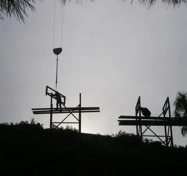 Trabajadores de la construcción silueteados construyen una estructura al aire libre, con una grúa levantando una viga.