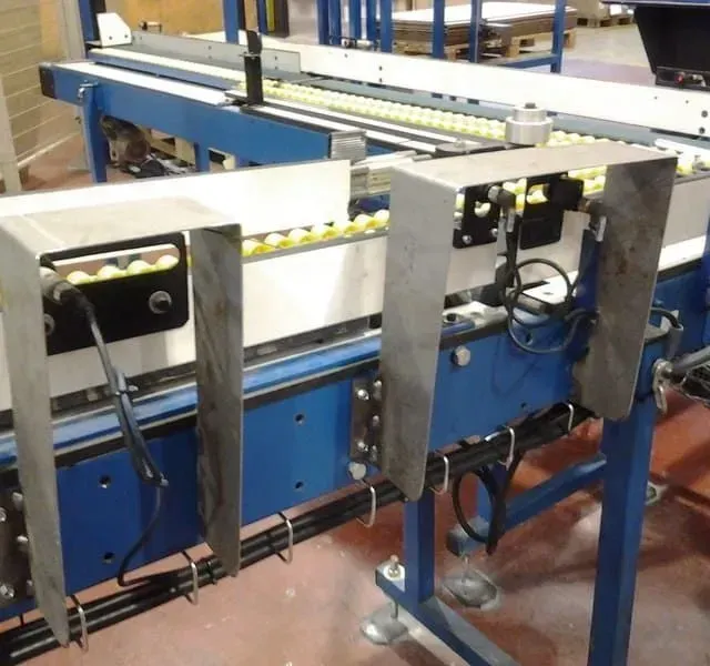 Sistema de cinta transportadora con protecciones metálicas, transportando objetos de color amarillo; marco azul.