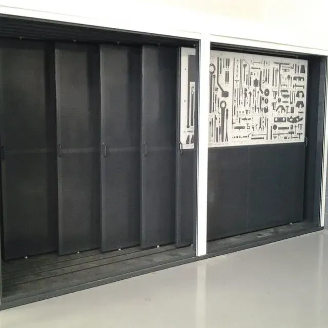 Paneles deslizantes gris oscuro que cubren una pared con un expositor de herramientas recortado. Marco blanco, suelo claro.