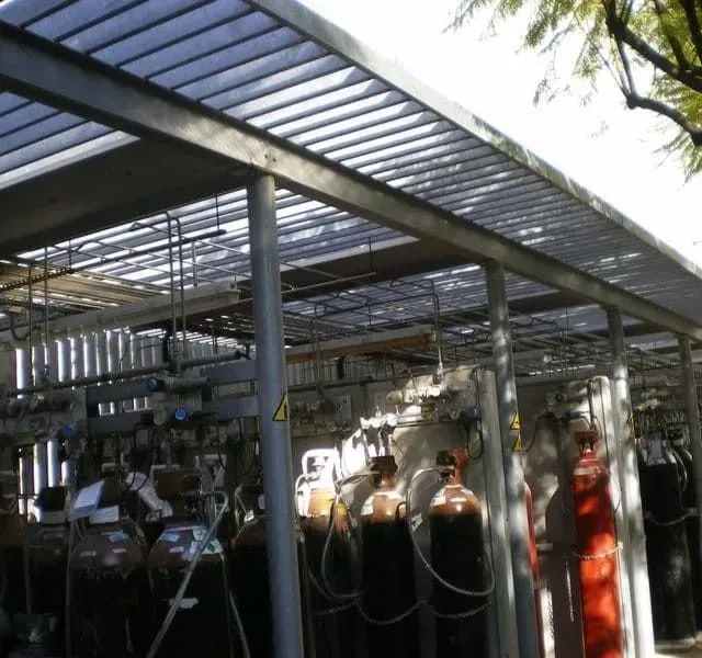 Almacenamiento de cilindros de gas al aire libre bajo techo metálico. Muchos cilindros son visibles, algunos rojos.