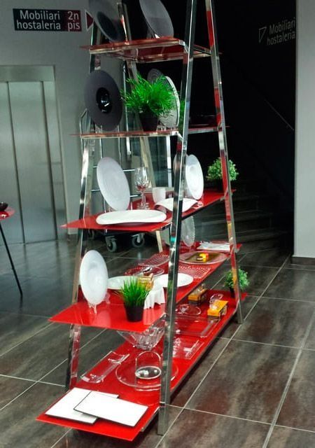 Estantería de exposición de color rojo y plata con platos, vasos y vegetación en una sala de exposición.