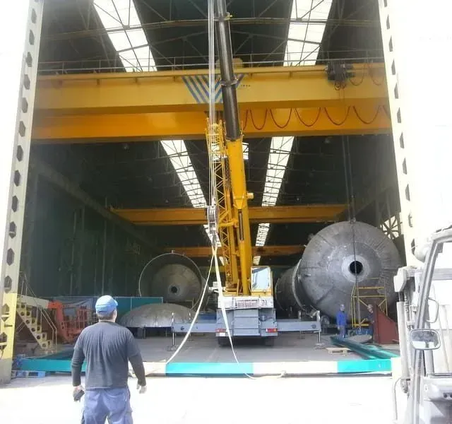 Una grúa en un almacén levanta tanques metálicos industriales. Un trabajador observa.