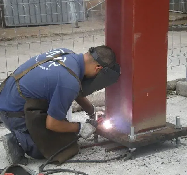 Soldador con equipo de protección soldando una base de columna de metal rojo sobre una superficie de hormigón al aire libre.