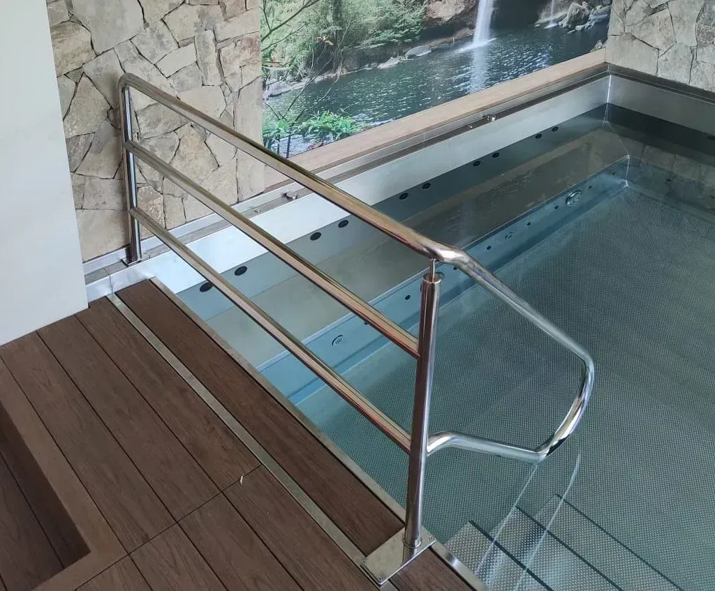 Piscina con pasamanos cromados, escalones y suelo imitación madera. Cascada en la pared detrás de la piscina.