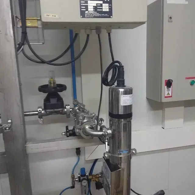 Tubería industrial de acero inoxidable con válvulas, sensor y panel de control eléctrico en una sala limpia.