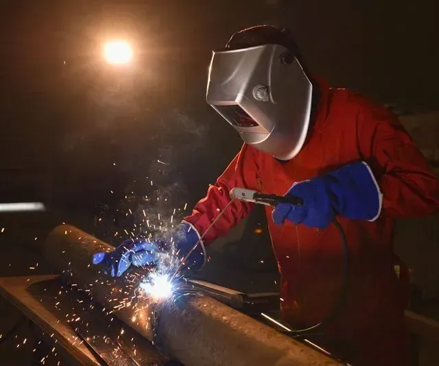 Soldador con mono rojo, soldando metal con chispas volando, con máscara protectora y guantes.
