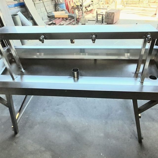 Mesa de acero inoxidable con tres grifos y desagüe central, probablemente para un entorno comercial.