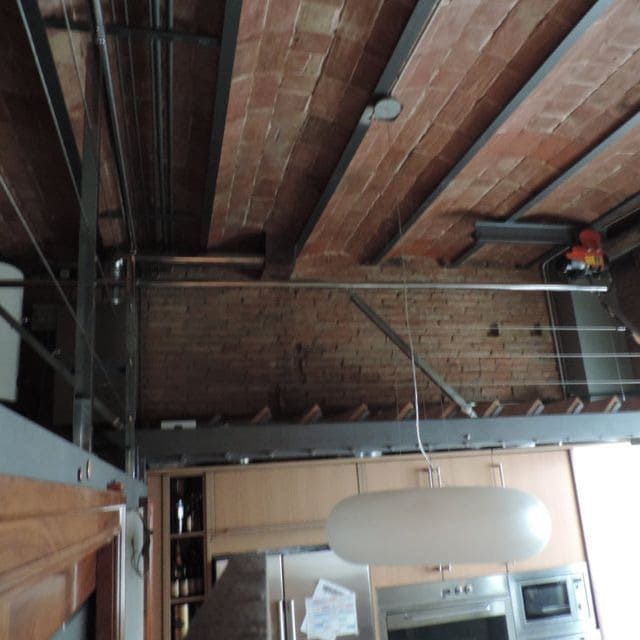 Interior con techo de ladrillo visto, vigas metálicas y armarios de cocina. Una lámpara cuelga.