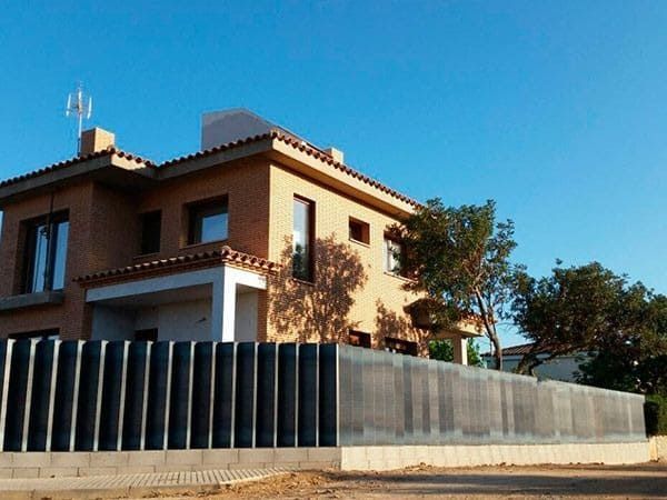 Casa de dos pisos de color canela con techo plano, detrás de una cerca de listones verticales negros, bajo un cielo azul.
