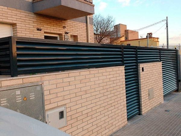 Muro de ladrillos rematado con una valla de listones negros y una puerta delante de un edificio.
