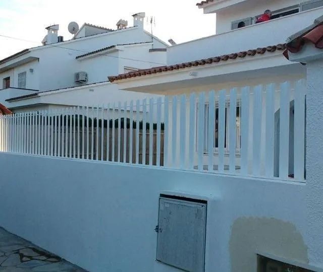 Valla blanca sobre una pared blanca, frente a un edificio blanco con techo de tejas rojas.