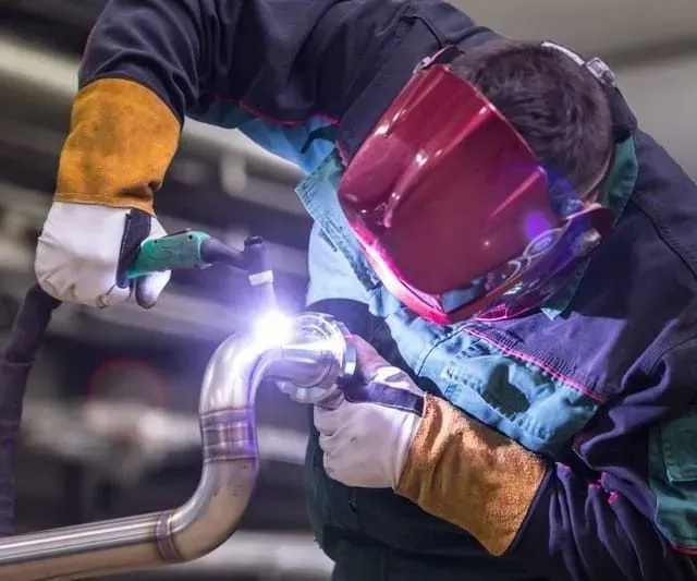 Soldador con equipo de protección utiliza un soplete sobre un tubo metálico curvo, creando una chispa brillante.