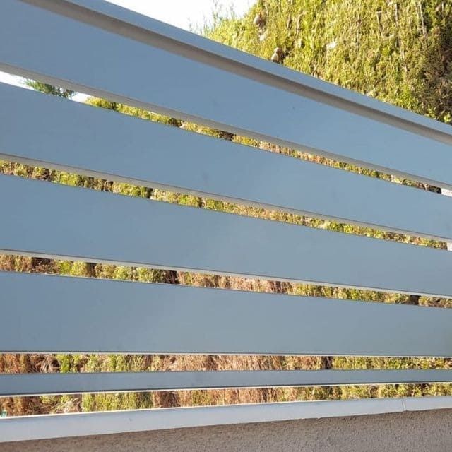 Valla de listones horizontales de color gris con espacios que revelan un fondo verde y marrón difuminado.