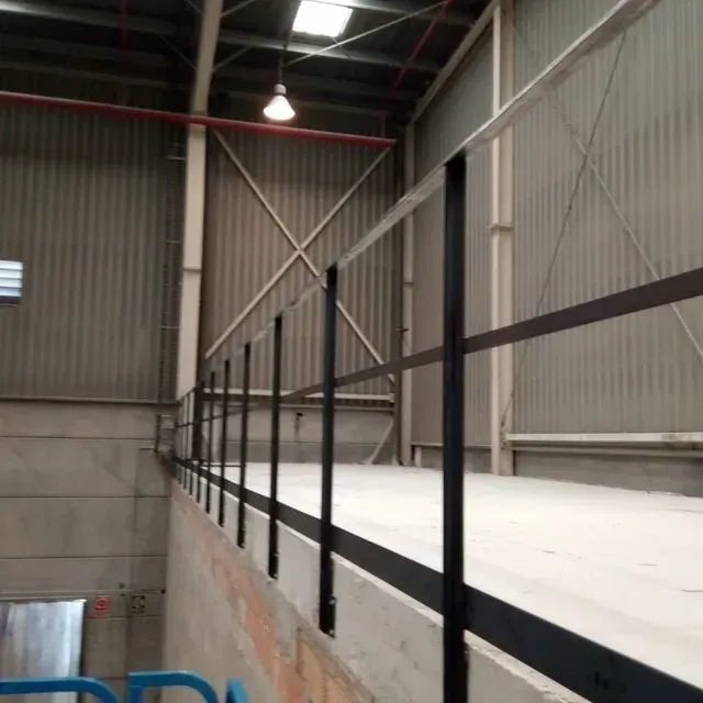 Barandilla negra a lo largo de una plataforma elevada dentro de un almacén; paredes de metal corrugado.