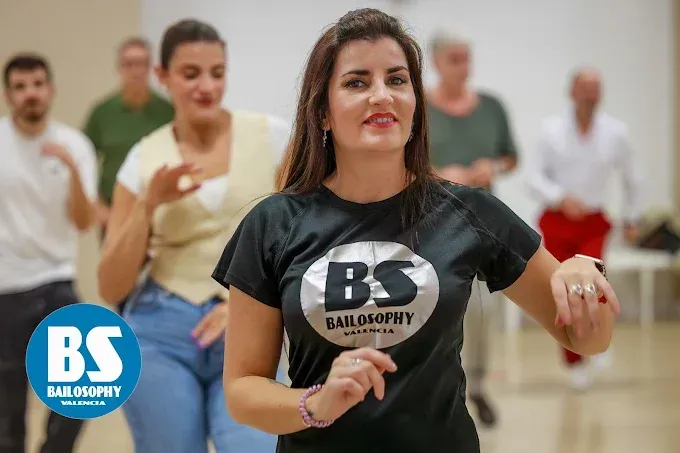Mujer con camiseta negra de baile de salón BS, bailando en un estudio con otras personas desenfocadas al fondo