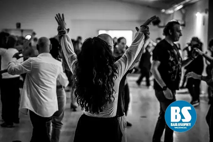 Personas bailando con los brazos en alto en un evento en interiores en blanco y negro, con un logotipo «BS» en la esquina