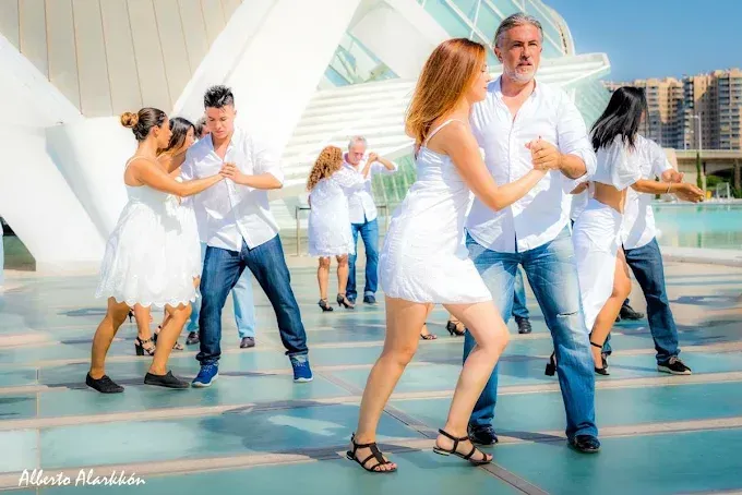 Personas bailando con ropa blanca en un paseo marítimo luminoso, con un puente moderno al fondo
