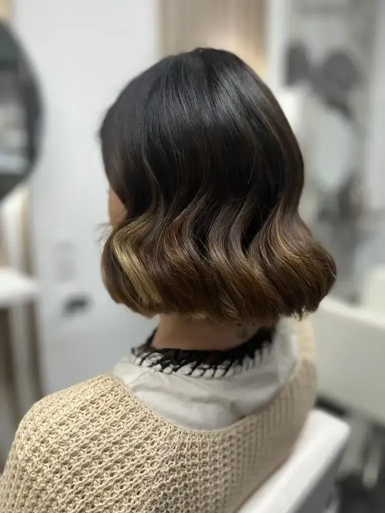 Vista posterior de un corte de pelo bob corto y ondulado con raíces oscuras y puntas rubias en una silla de peluquería.