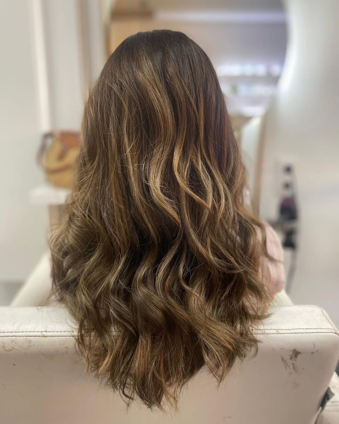 Cabello largo y ondulado castaño desde atrás, sentada en una silla blanca en una habitación tenuemente iluminada.