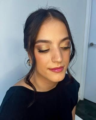 Primer plano de una mujer de cabello oscuro vestida de negro, con maquillaje dorado en los ojos, mirando hacia abajo, frente a una pared interior lisa.