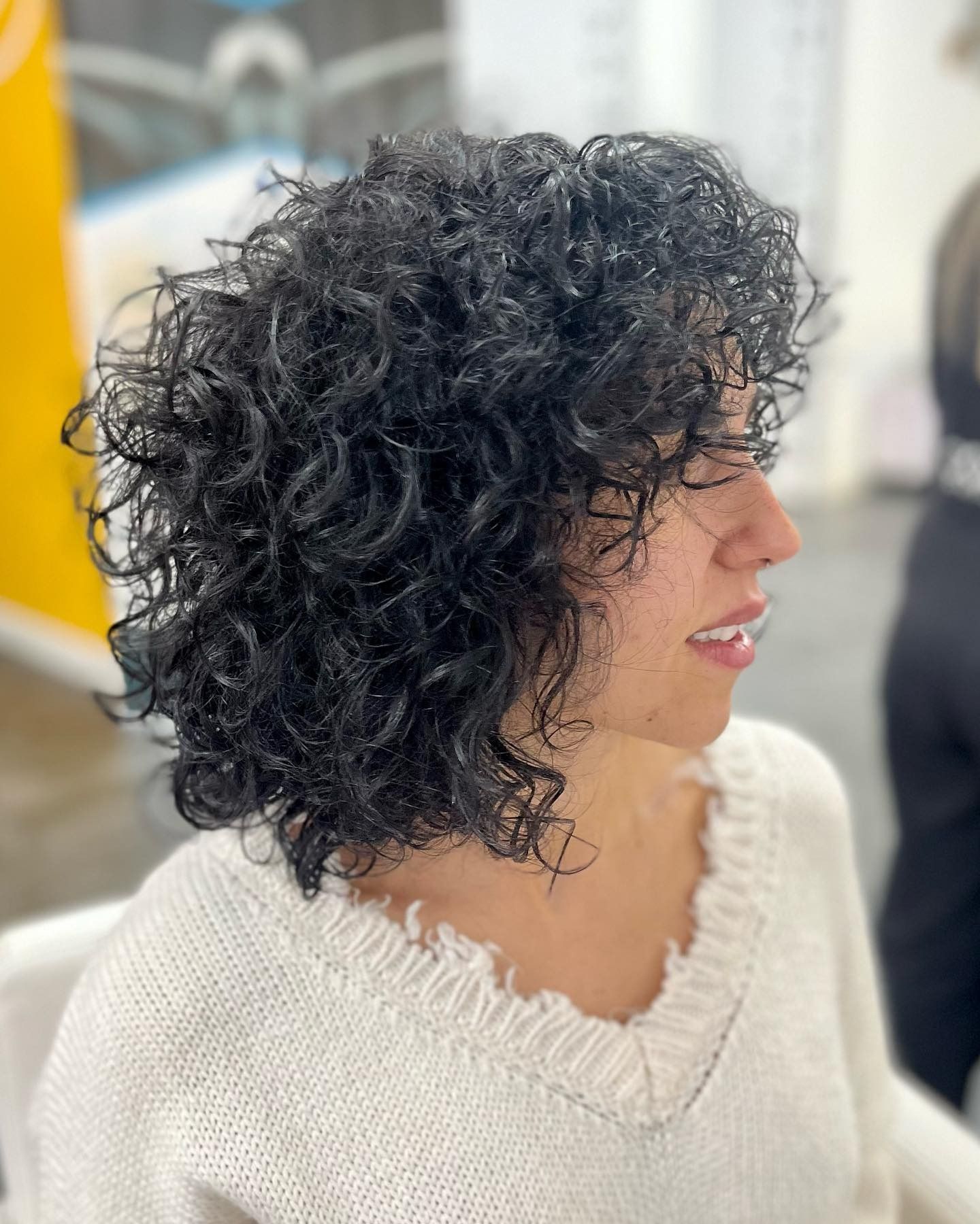 Persona de cabello rizado con un suéter color crema, vista de perfil en interiores, sonriendo levemente.