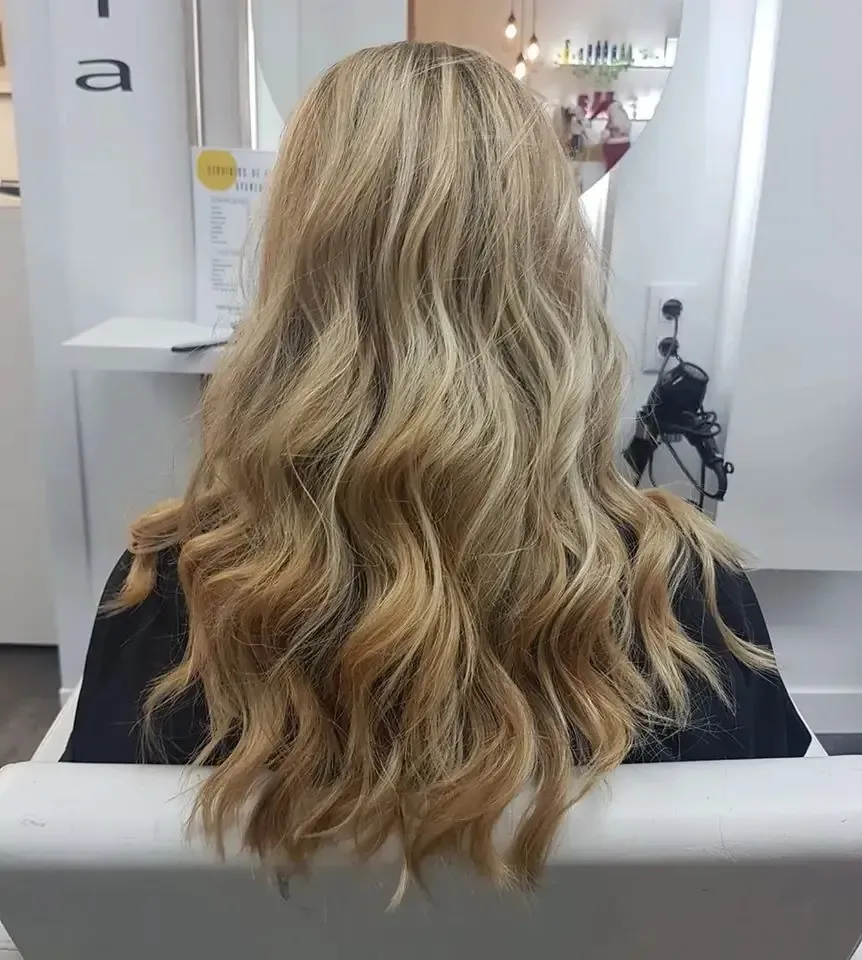 Cabello rubio ondulado visto desde atrás en una silla de peluquería, con un espejo y herramientas de peinado delante.