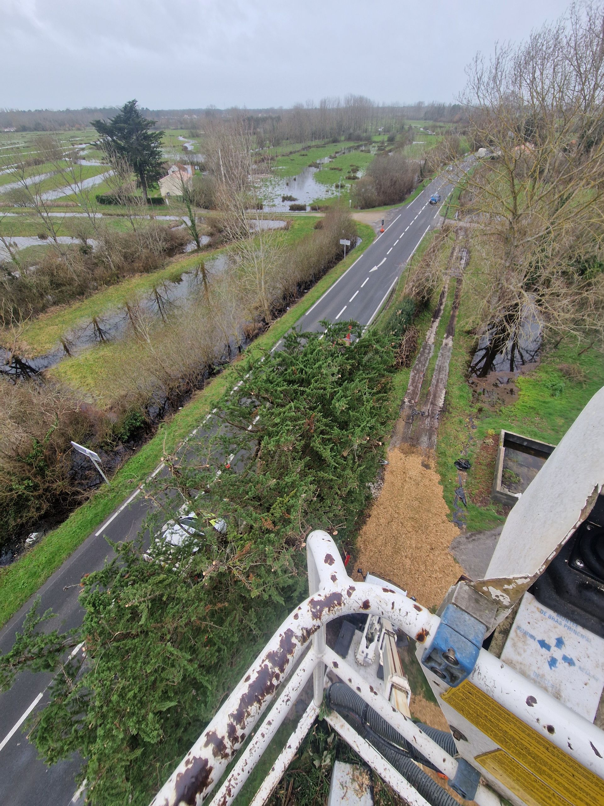 Vue depuis une nacelle élévatrice surplombant une route bordée d'arbres, d'un bas-côté herbeux et d'un petit fossé.