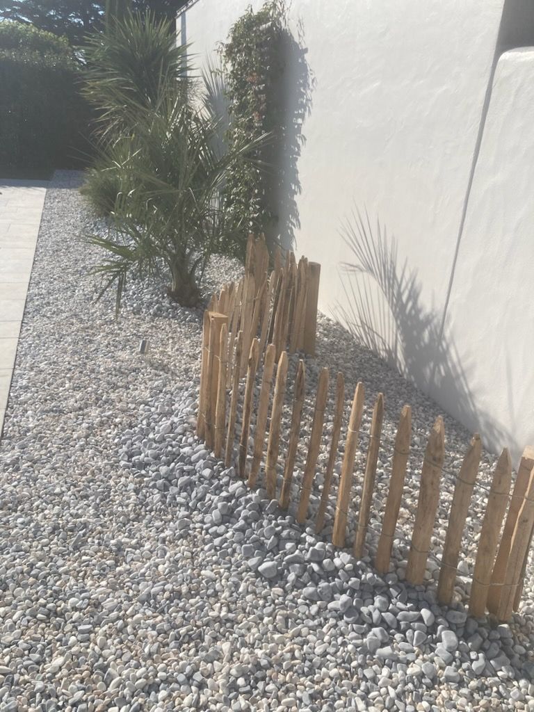 Une petite clôture en piquets de bois incurvés repose sur un lit de gravier gris clair, à côté d'une plante ressemblant à un palmier et d'un mur blanc.