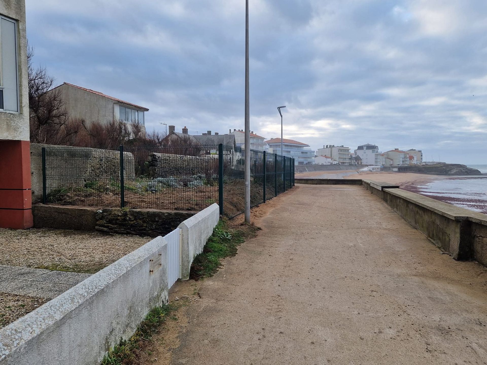 Un chemin de gravier longe un muret en pierre, à côté d'un jardin clôturé, avec des maisons en arrière-plan, au bord de la mer.