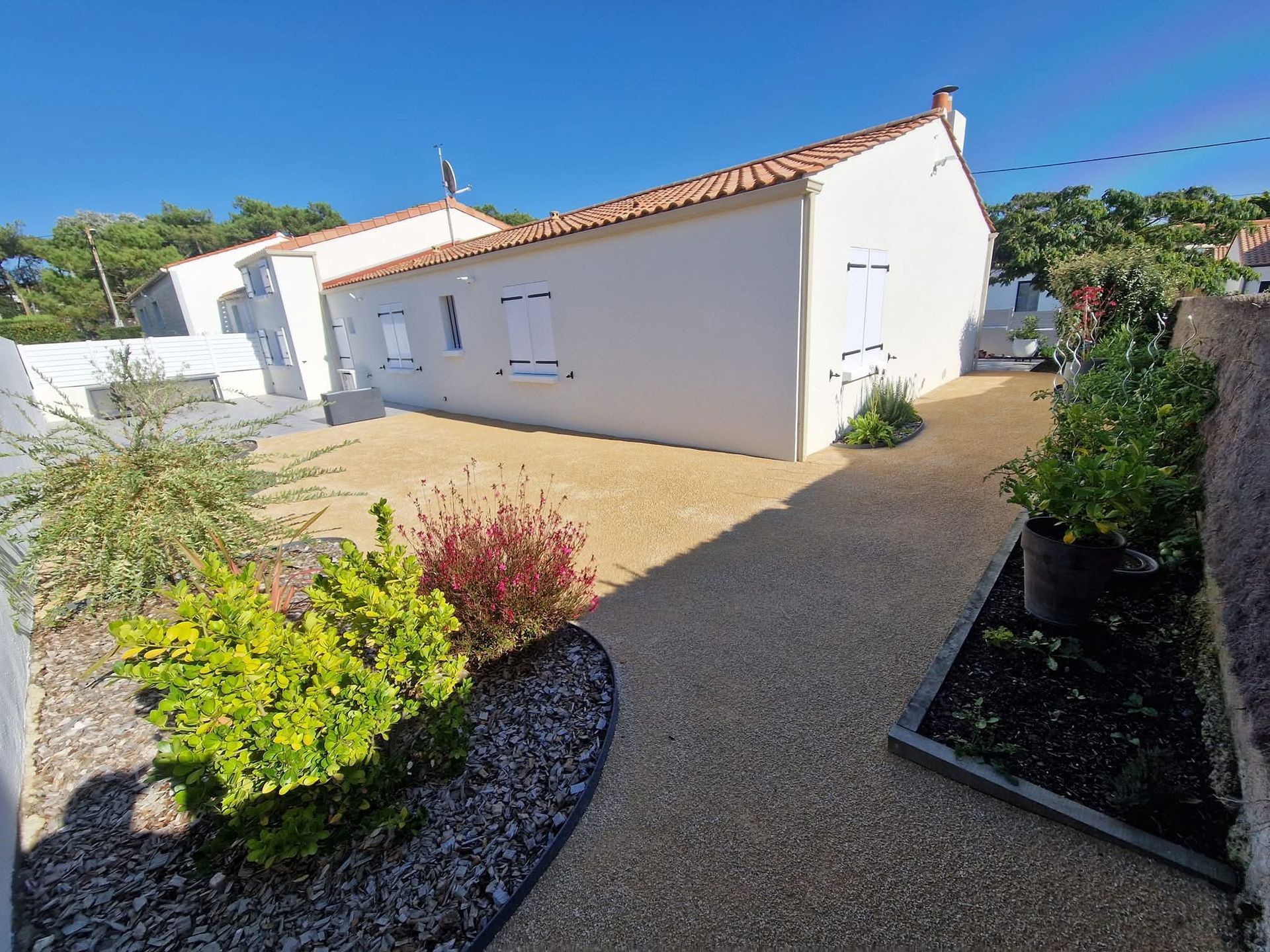 Une maison blanche au toit de tuiles surplombe une cour de gravier agrémentée de parterres de fleurs, vue par une journée ensoleillée.