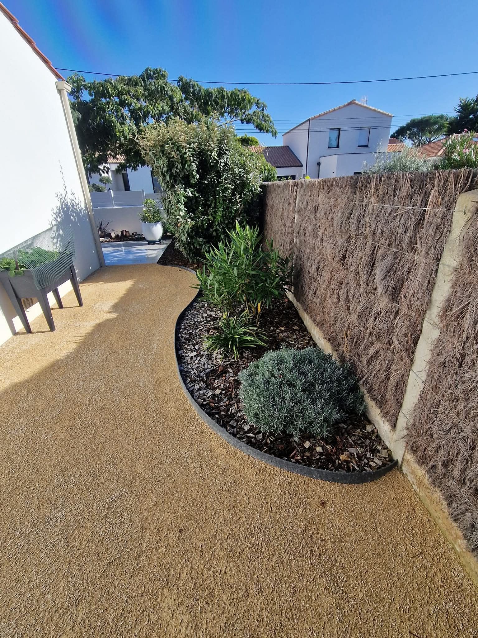 Un chemin de gravier sinueux longe un petit parterre de fleurs avec des arbustes, adossé à un mur de pierre texturé, à côté d'une maison blanche.