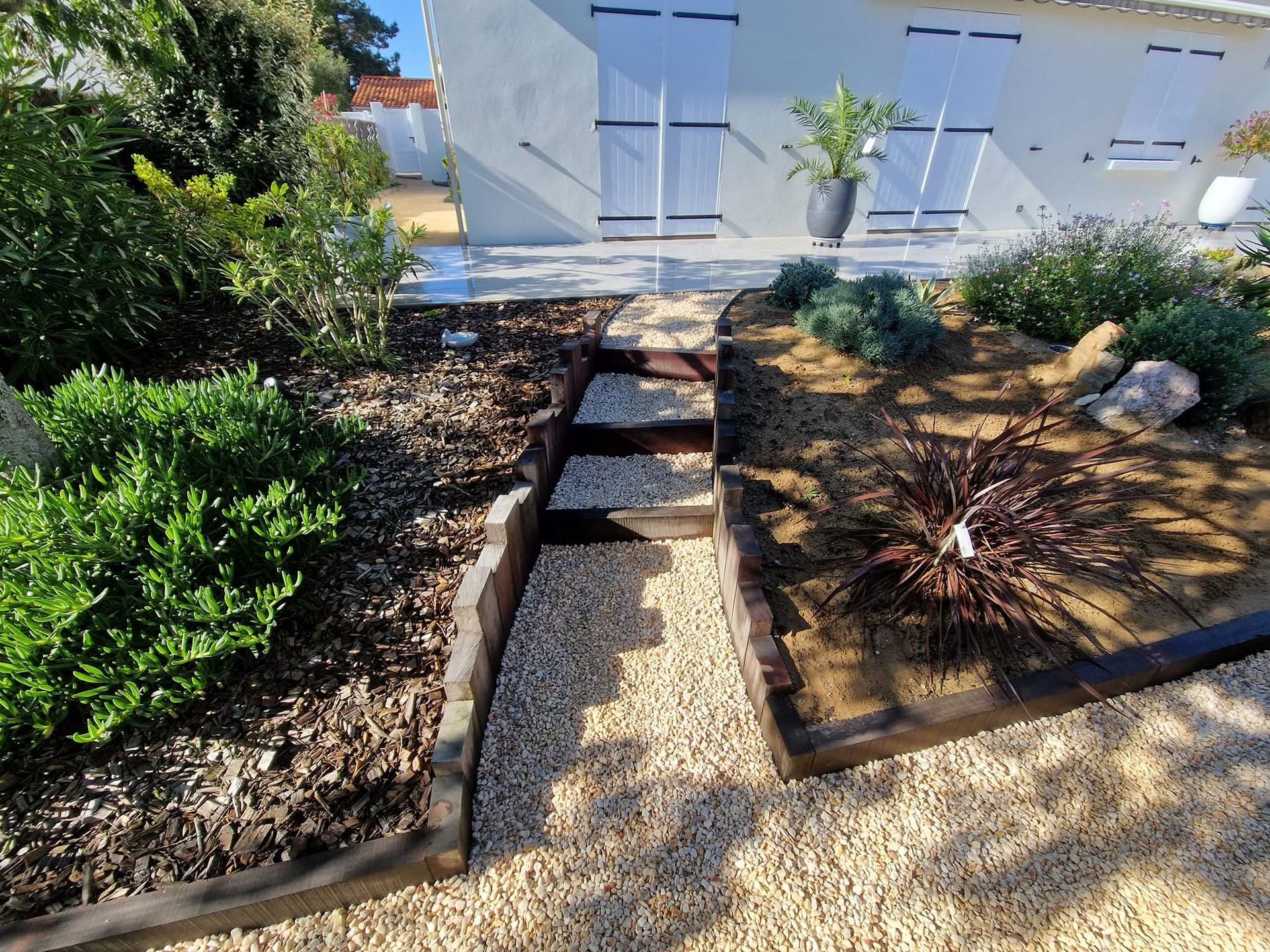 Un chemin de gravier avec des marches en bois mène à une maison blanche aux volets blancs, entourée de parterres de fleurs.
