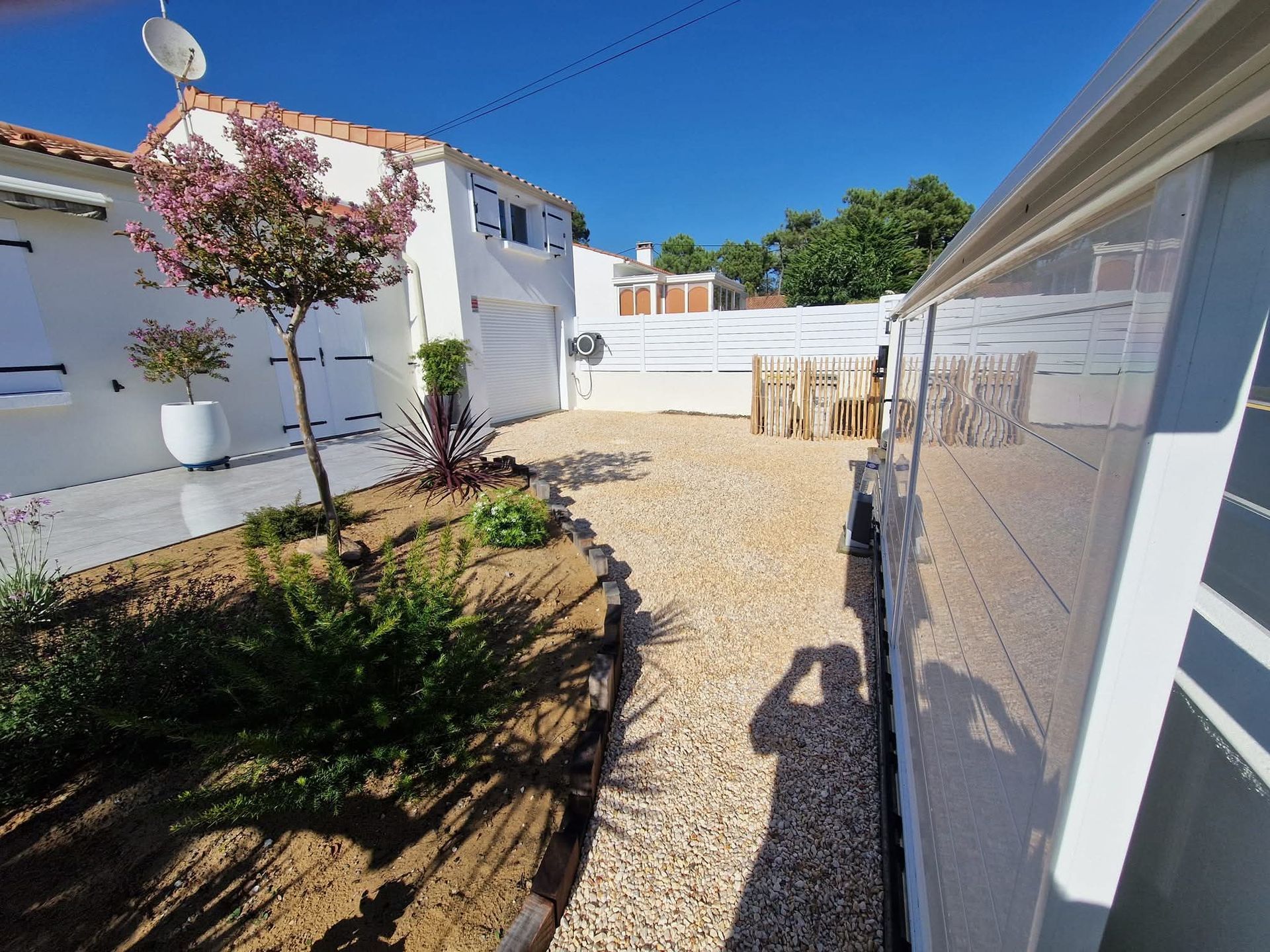 Une maison blanche avec une cour gravillonnée, un petit parterre de jardin aménagé et une structure de patio couverte partiellement visible.