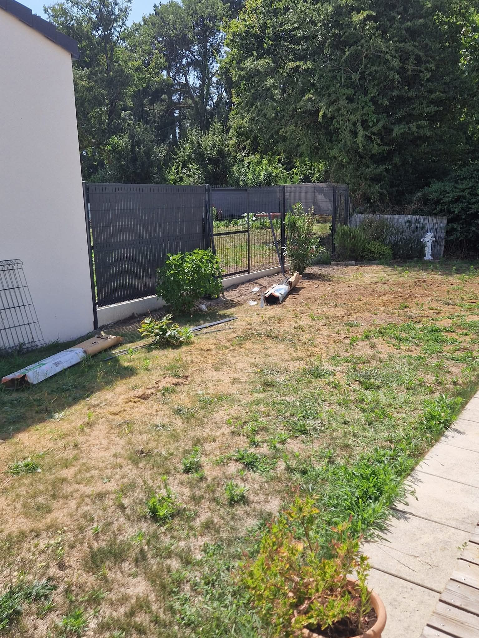 Une cour latérale avec un mur blanc, une clôture grise, des matériaux de construction éparpillés et une pelouse sèche et clairsemée.