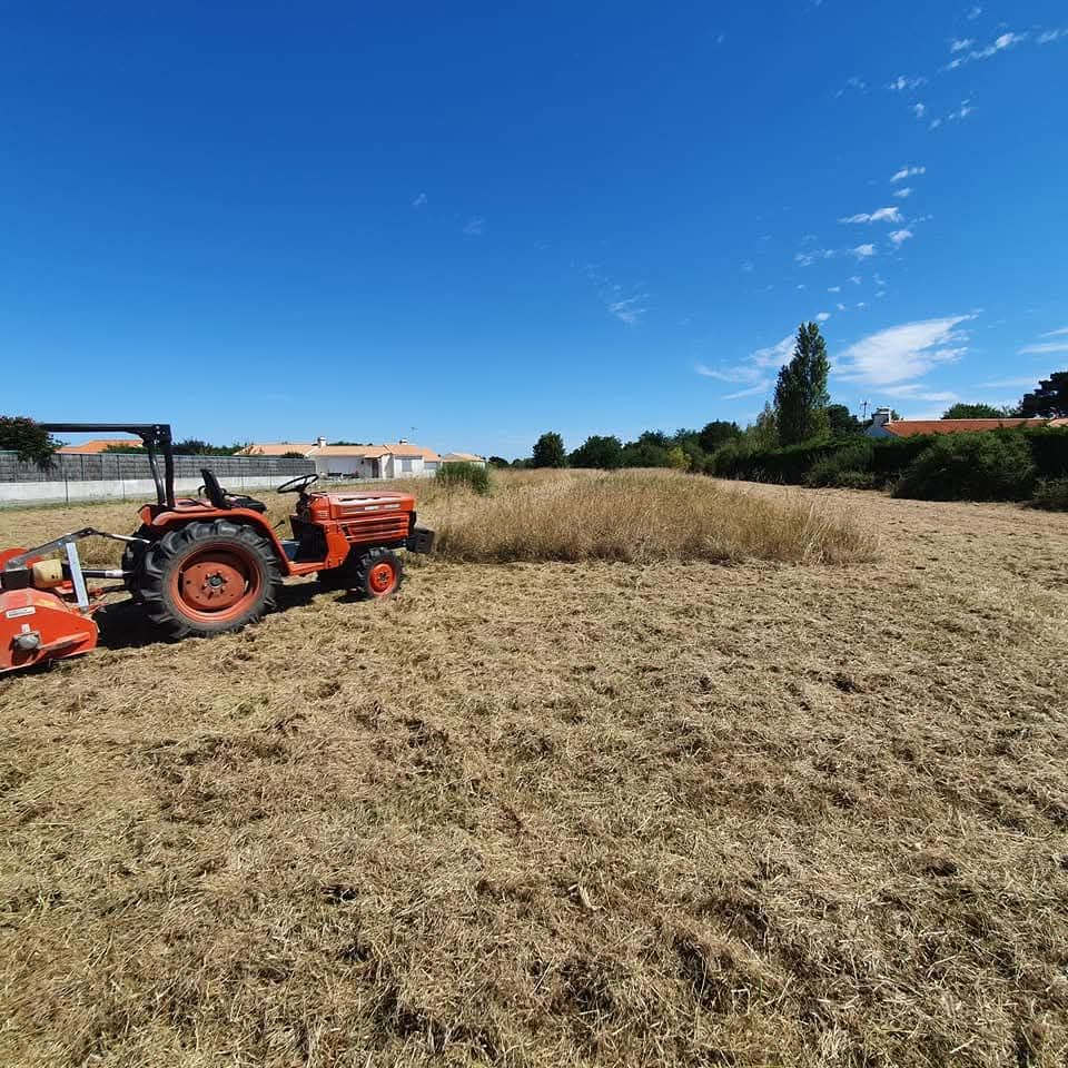 Un tracteur orange équipé d'une fraise est stationné dans un champ sec et fauché, par une journée ensoleillée sous un ciel bleu dégagé.