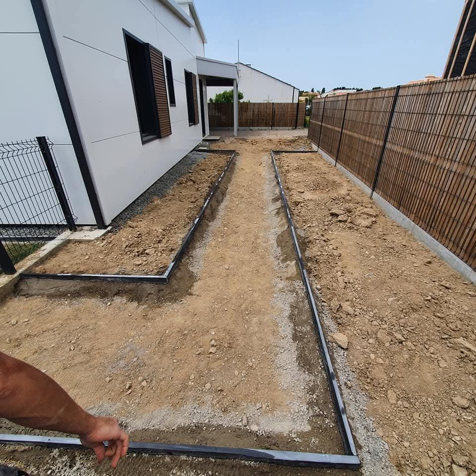 Un étroit chemin de terre en construction entre une maison moderne blanche et une clôture en lattes de bois, encadrée par des bordures métalliques.
