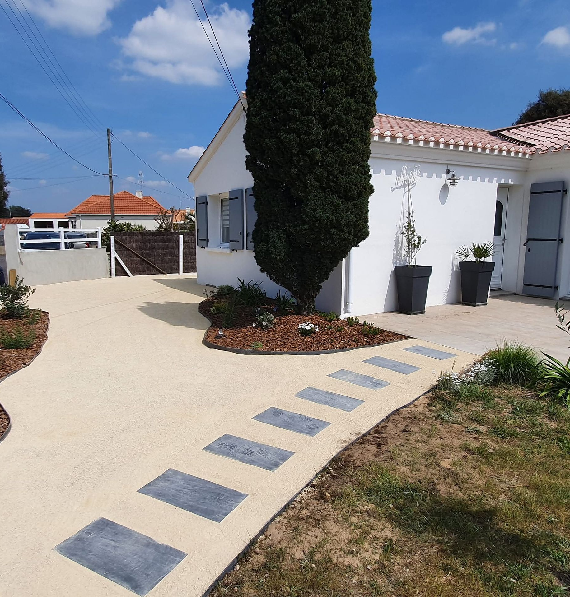 Une maison blanche avec un grand cyprès et une allée beige pavée de dalles rectangulaires grises.