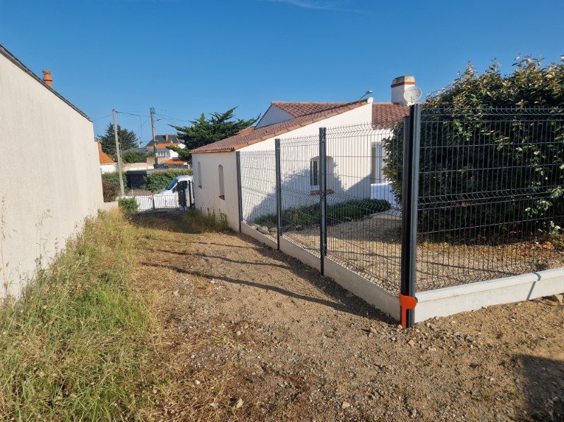 Un chemin de terre longe une maison blanche au toit de tuiles rouges, séparée par une clôture métallique noire sur une base en béton.