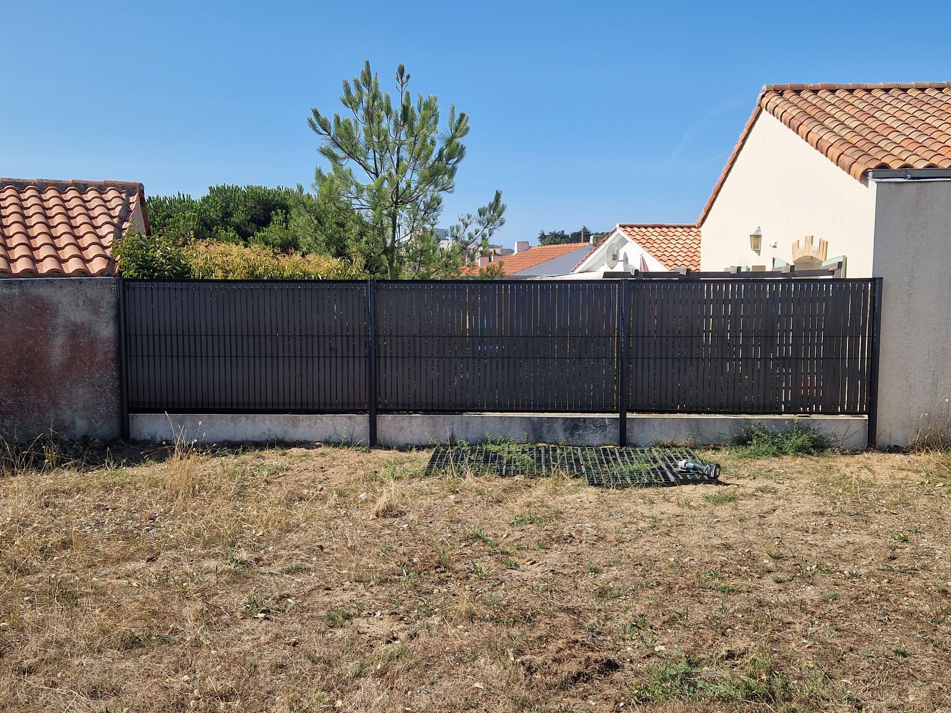 Une clôture métallique sombre, empreinte de confidentialité, repose sur une base en béton basse, dans une cour à l'herbe sèche et brune, sous un ciel d'un bleu limpide.