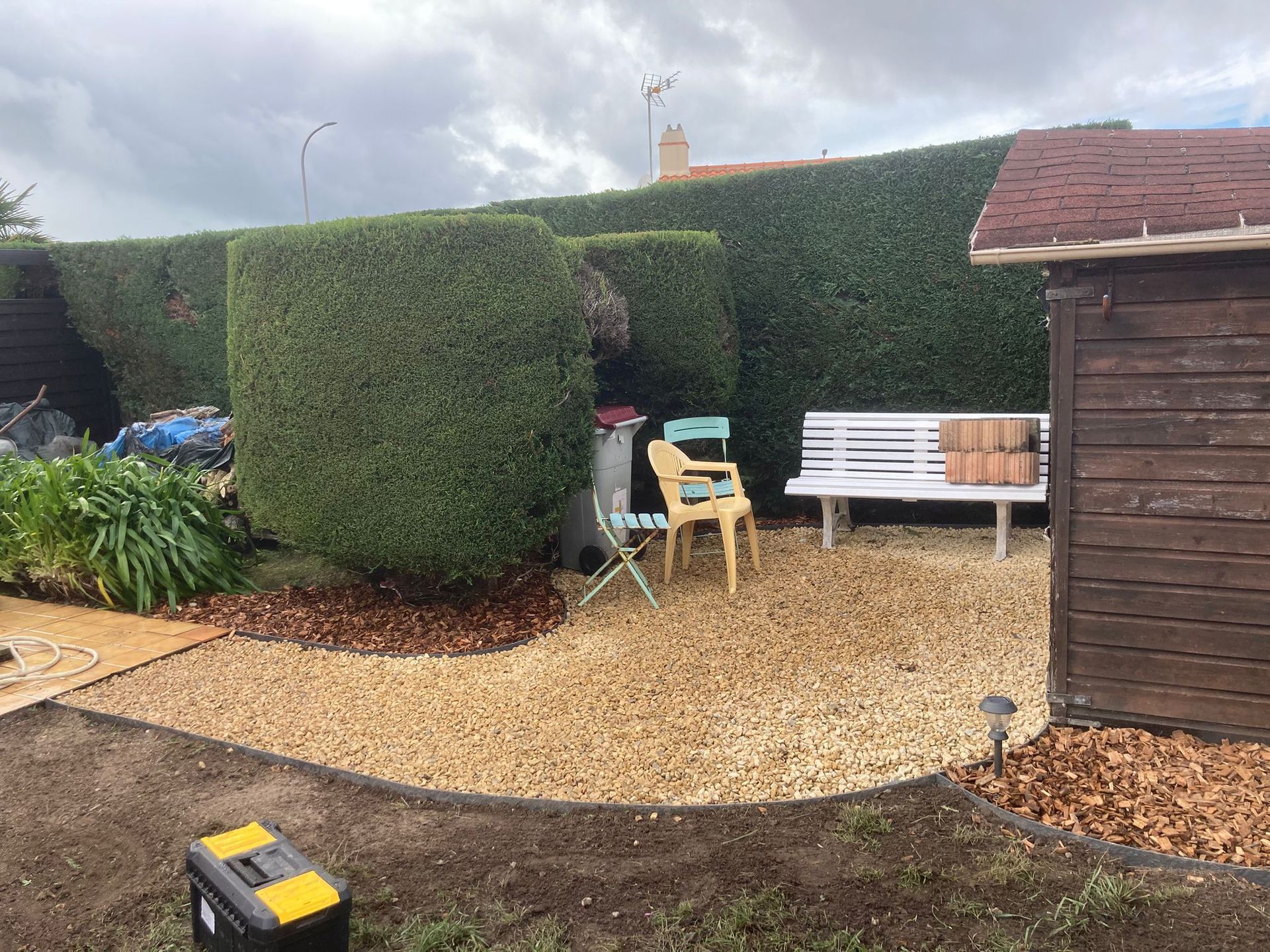 Une scène de jardin avec un coin salon en gravier, un banc blanc, une chaise jaune, une grande haie et un abri de jardin en bois foncé.