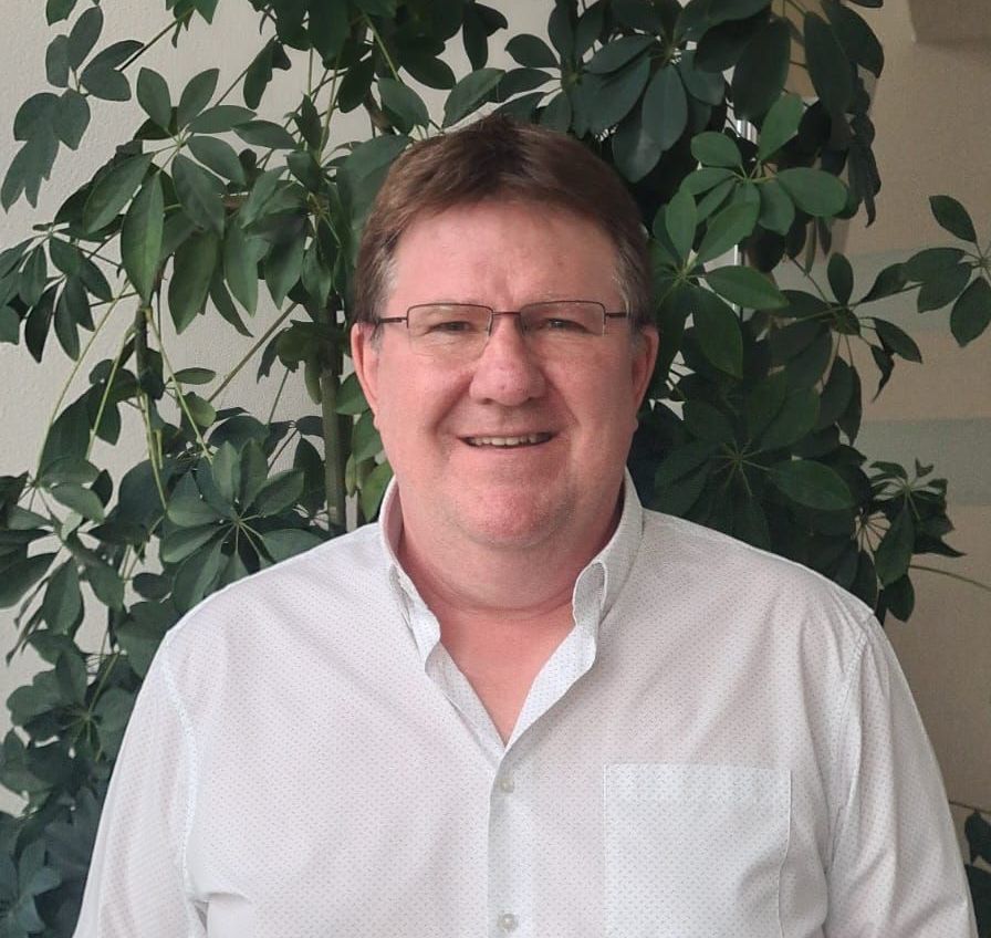 Ein Mann mit Brille und weißem Hemd lächelt vor einer Pflanze