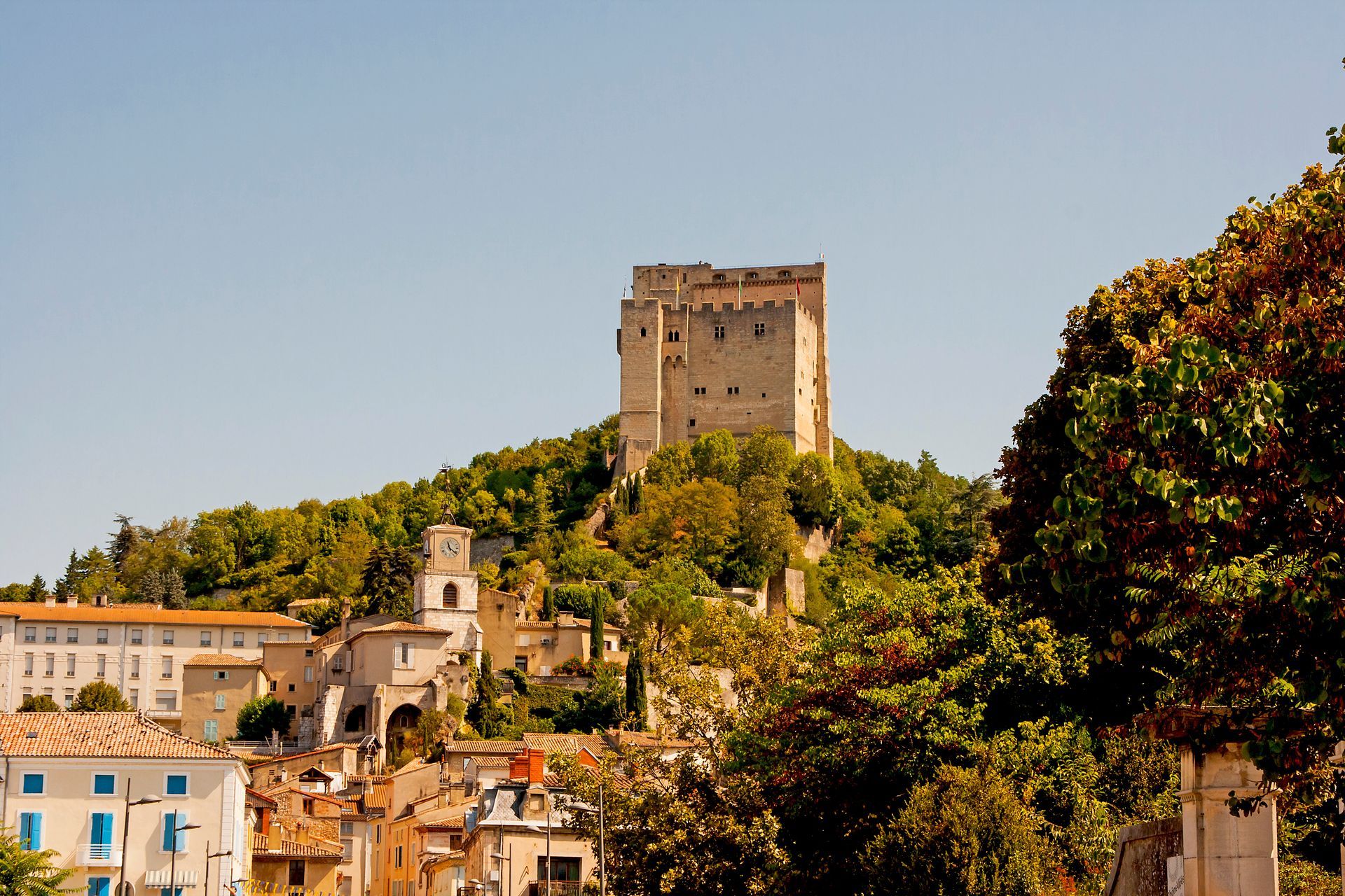 Château de pierre perché sur une colline, surplombant des bâtiments et des arbres colorés sous un ciel d'un bleu limpide