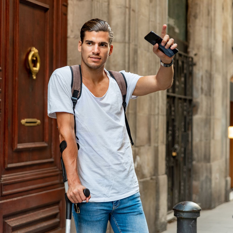 Voyageur tenant un téléphone et un petit appareil, debout dans la rue avec un sac à dos et une valise à roulettes.